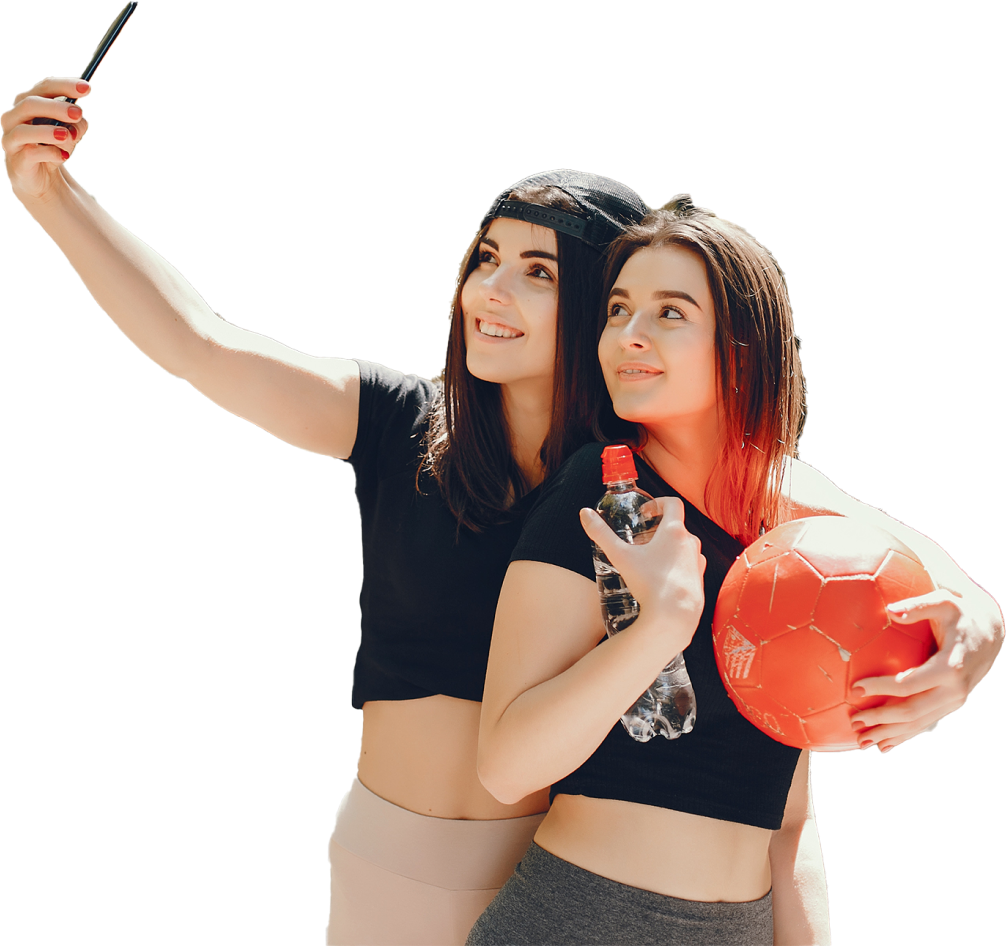 Two young women in sportswear, one holding a soccer ball and the other taking a selfie, standing outdoors under sunlight.